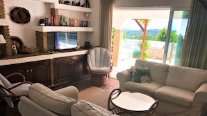 Uma sala de estar com sofá, poltronas, uma mesinha, televisão e estante.