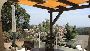Um pátio externo com mesa e cadeiras de madeira, um banco e uma pérgola com toldo amarelo.