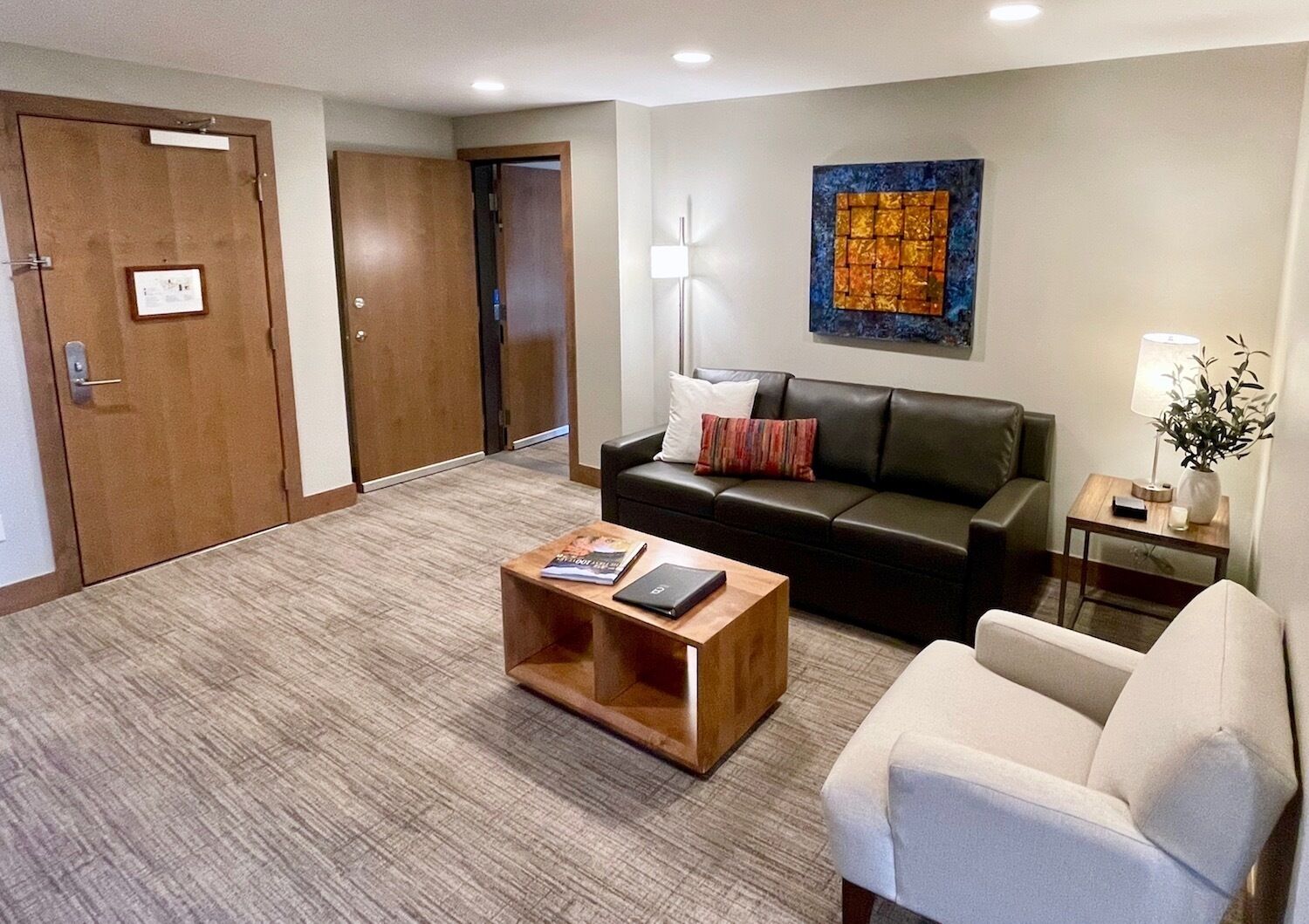 A hotel lobby with a leather sofa, a wooden coffee table, a white armchair, a small side table with a lamp, and a painting on the wall.