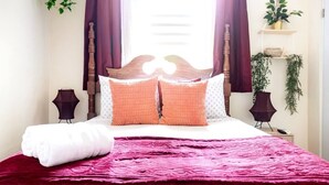 Un lit bien fait, recouvert d’une couverture rouge, avec deux oreillers orange et un oreiller blanc. On y trouve deux lampes de chevet, une petite étagère avec une plante et une fenêtre avec des rideaux.