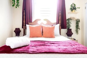 A neatly made bed with a red blanket, two orange pillows, and a white pillow. There are two bedside lamps, a small shelf with a plant, and a window with curtains.