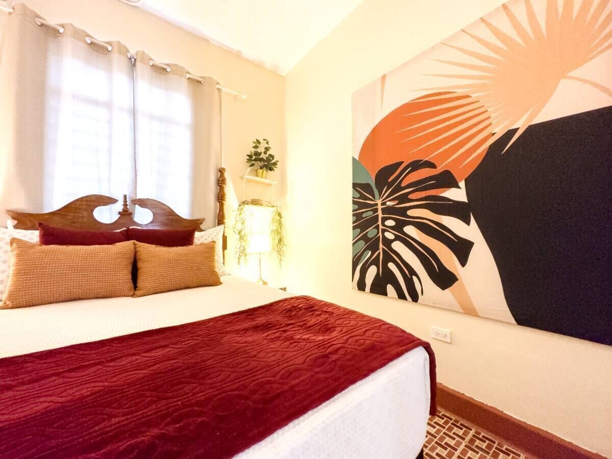 A hotel room with a large bed, a window with curtains, a wall-mounted lamp, and a large abstract painting.