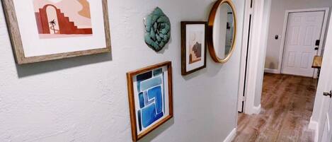 A hallway with a light gray wall, wooden flooring, and several framed artworks including a landscape, abstract, and geometric designs.