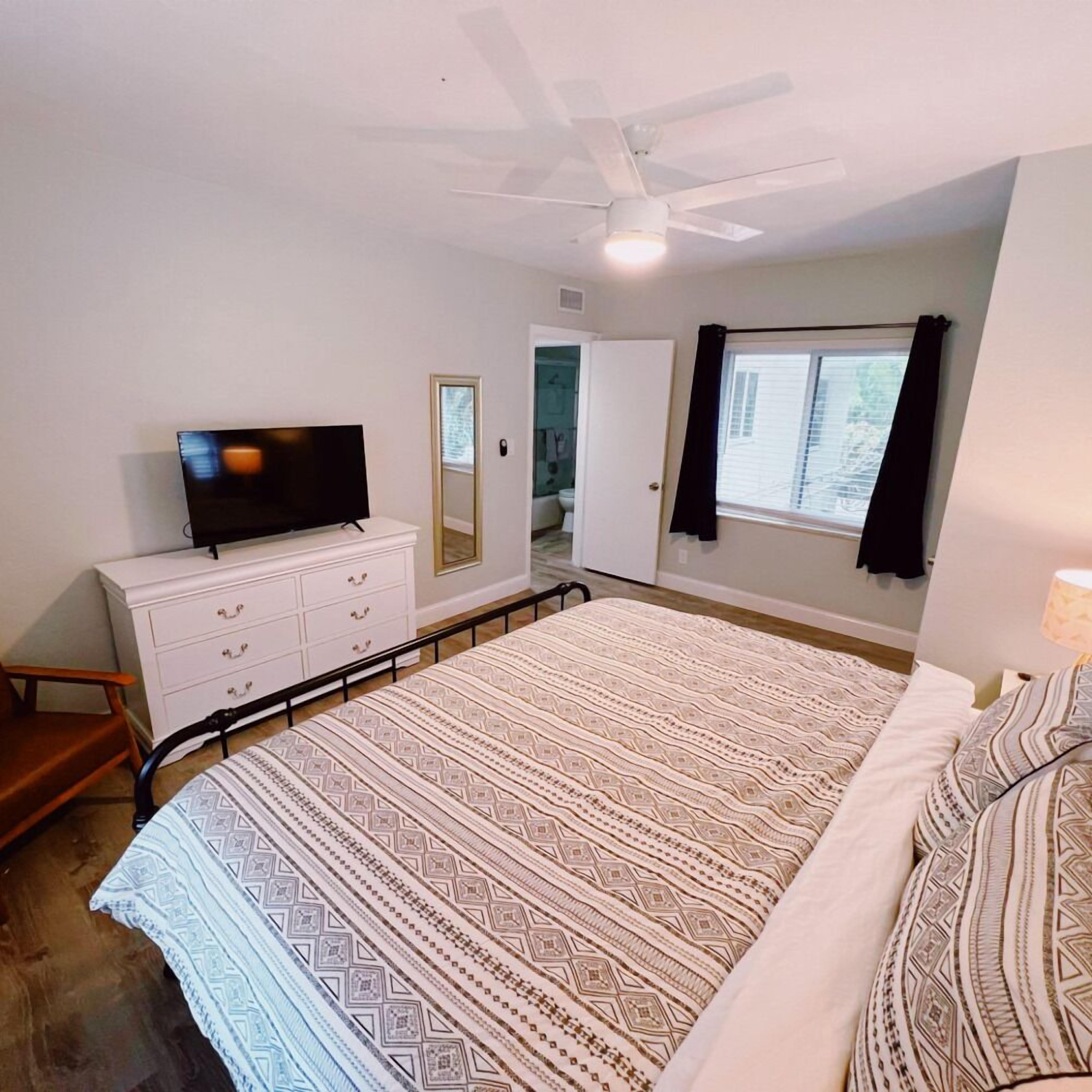 A bedroom with a bed, a TV on a dresser, a ceiling fan, a window with curtains, and a door leading to another room.