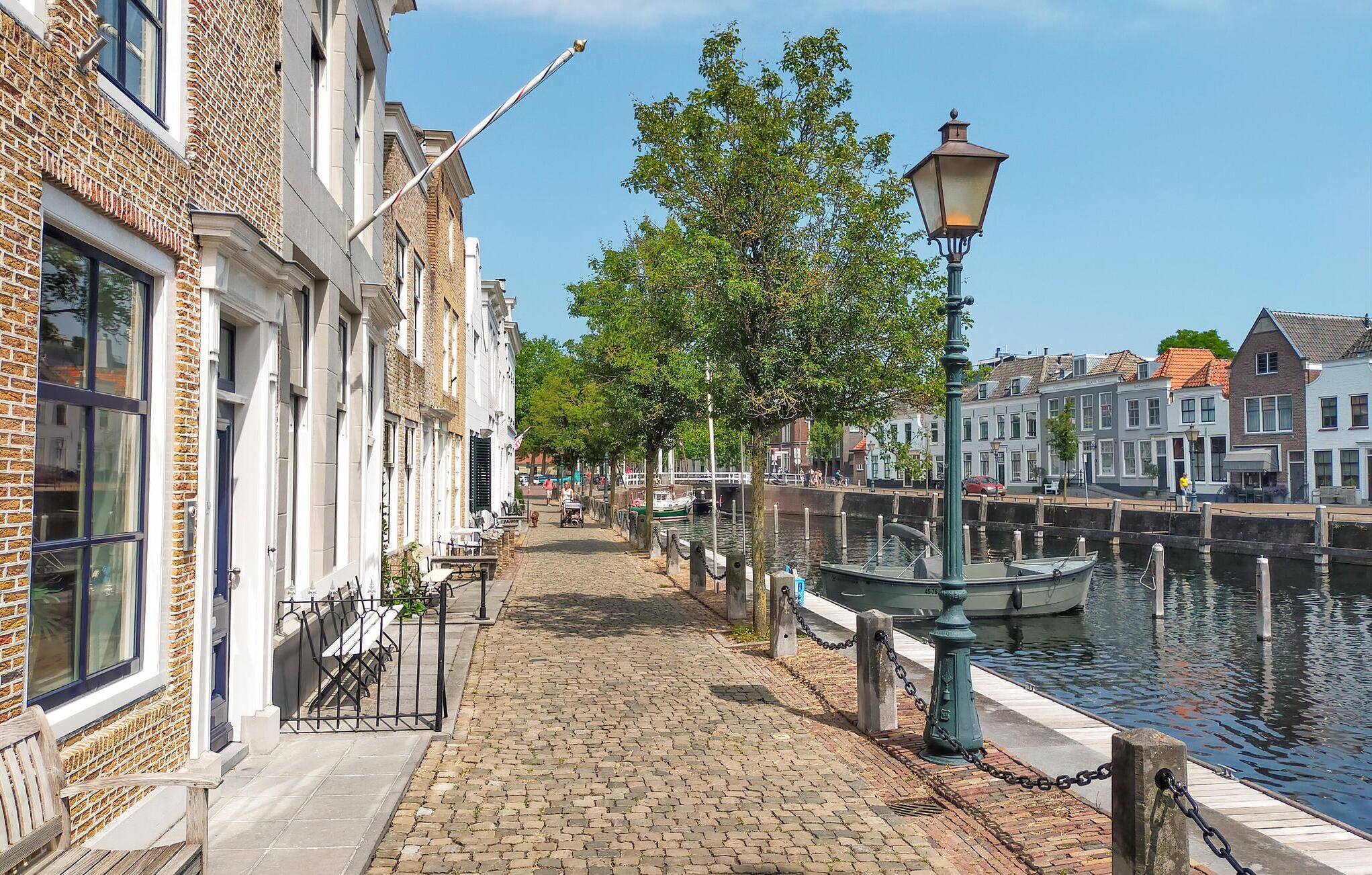 Een geplaveid pad langs een kanaal, met aan de ene kant traditionele gebouwen en aan de andere kant een rij aanmerende boten.