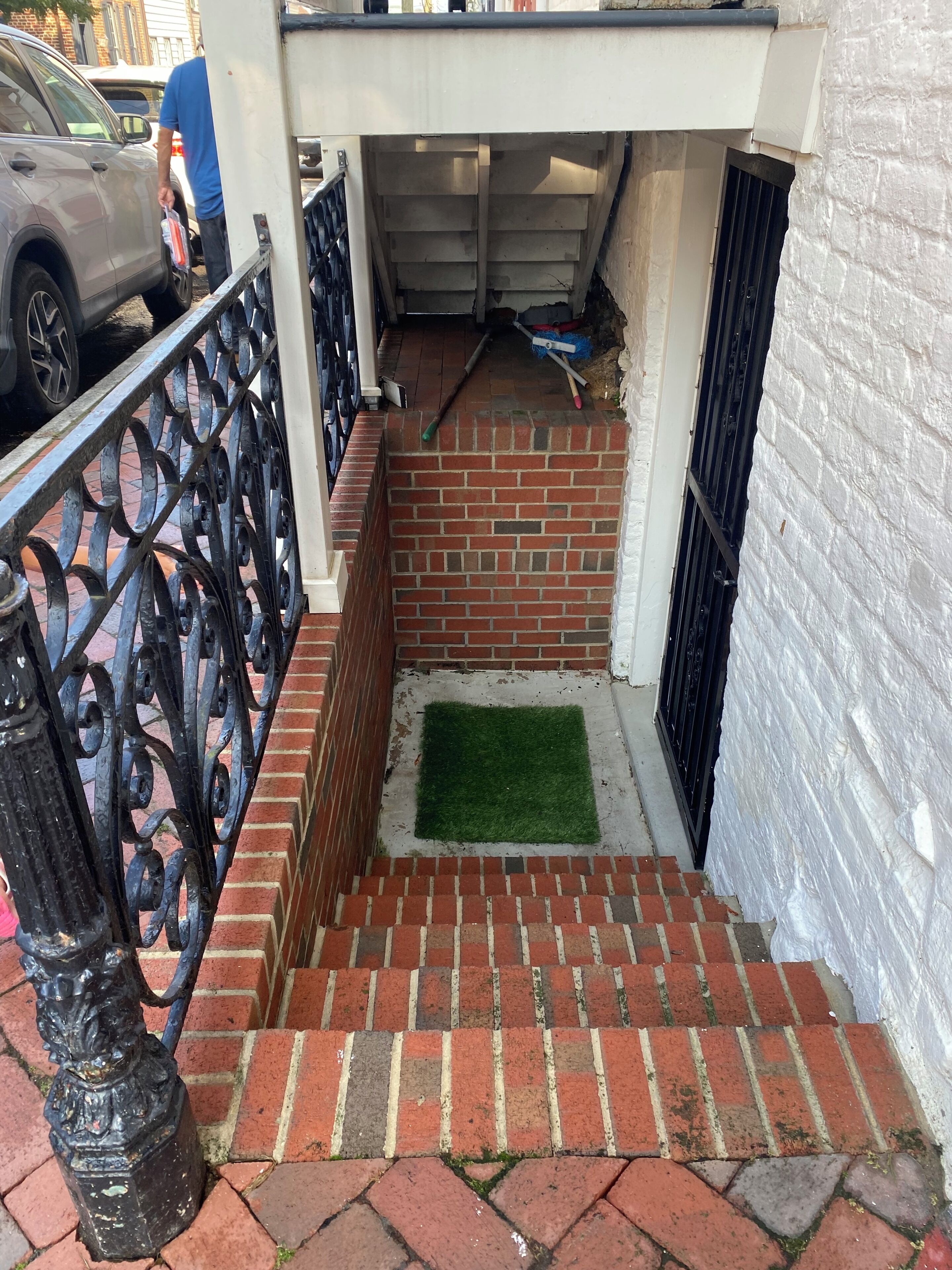Uma escadaria de tijolos estreita leva a uma entrada coberta, com um corrimão de metal de um lado e uma parede branca do outro.