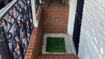 Uma escadaria de tijolos estreita leva a uma entrada coberta, com um corrimão de metal de um lado e uma parede branca do outro.
