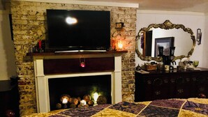 Una habitación con chimenea de ladrillo, un televisor montado encima, un espejo con marco decorativo y una cama con colcha.