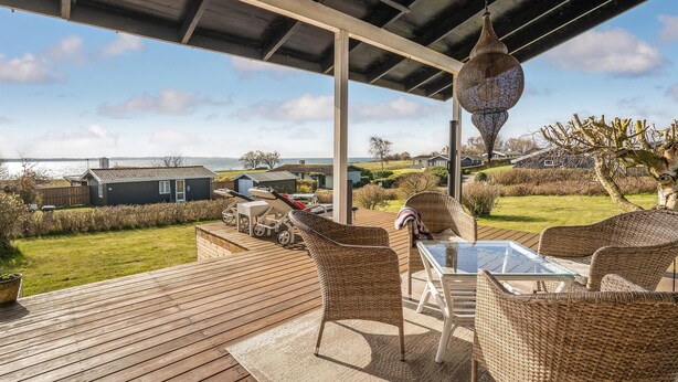Una terraza de madera con mobiliario de mimbre y una mesa de vidrio, con vistas a un paisaje costero con casas y una extensión de agua.