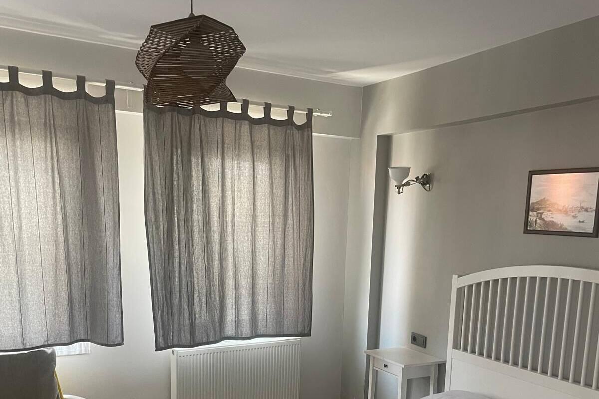 Quarto com cama, cadeira, mesa de cabeceira, luminária e janela com cortinas.