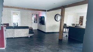 Un salón amplio con suelo oscuro, un conjunto de sofás, un televisor montado en la pared y una cocina con taburetes de bar.