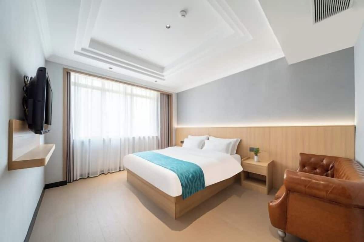 A hotel room with a bed, a brown leather armchair, a bedside table with a lamp, and a TV mounted on the wall.