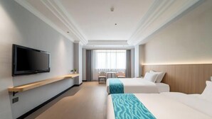 A hotel room with a flat-screen TV mounted on the wall, a bed with a blue patterned bedspread, a bedside table, and a window with curtains. - Xilai Hotel (Yueqing)