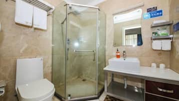 Un baño con ducha envuelta en vidrio, inodoro blanco, lavamanos con artículos de aseo y un espejo.