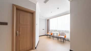 A hotel room with a wooden door, a small table with chairs, a large window with curtains, and a tiled floor.