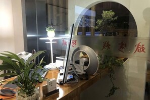 Una mesa de recepción con computadora, teléfono y una planta. Se ve una ventana con vistas al exterior.