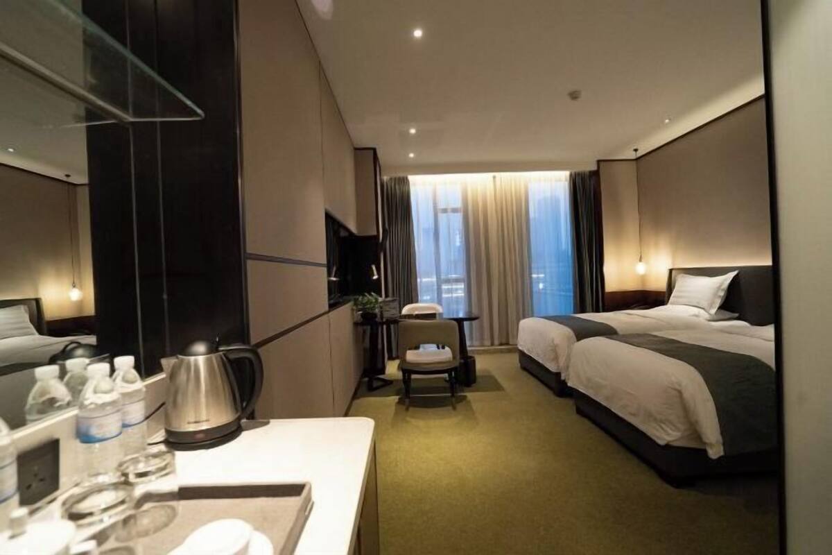 A hotel room with a bed, a desk with a kettle, a chair, and a small table with water bottles.