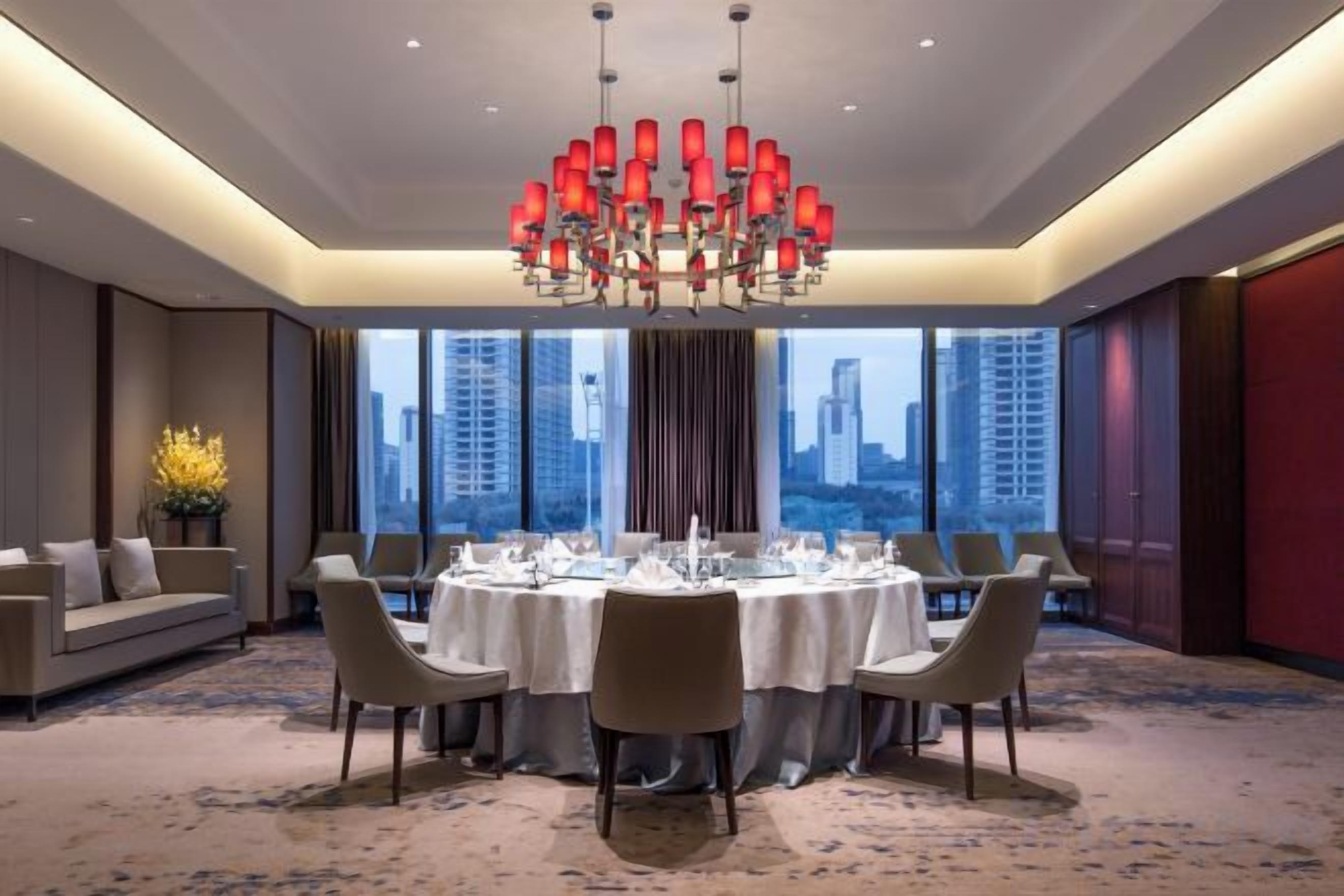 A hotel dining room with a round table set for six, large windows, and a chandelier.
