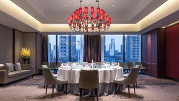 A hotel dining room with a round table set for six, large windows, and a chandelier.