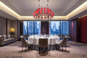 A hotel dining room with a round table set for six, large windows, and a chandelier.