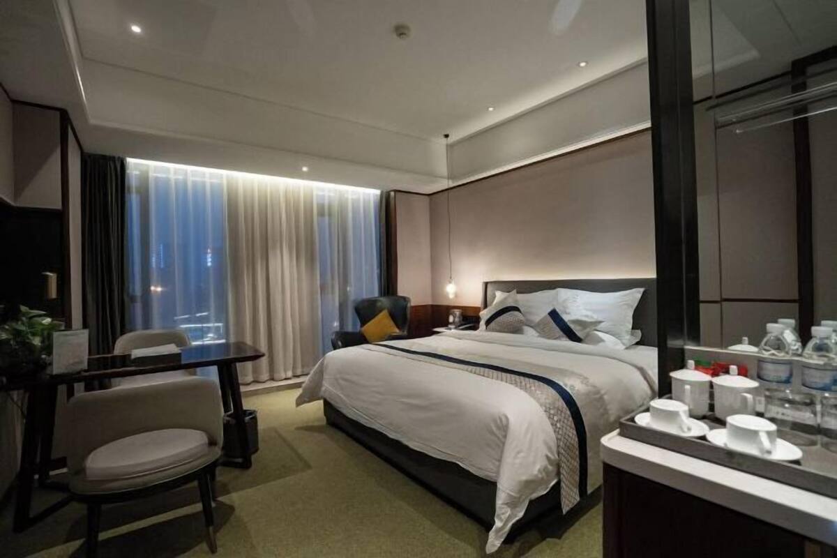 A hotel room with a large bed, a desk, a chair, a bedside table with cups, and a view of the city through the window.