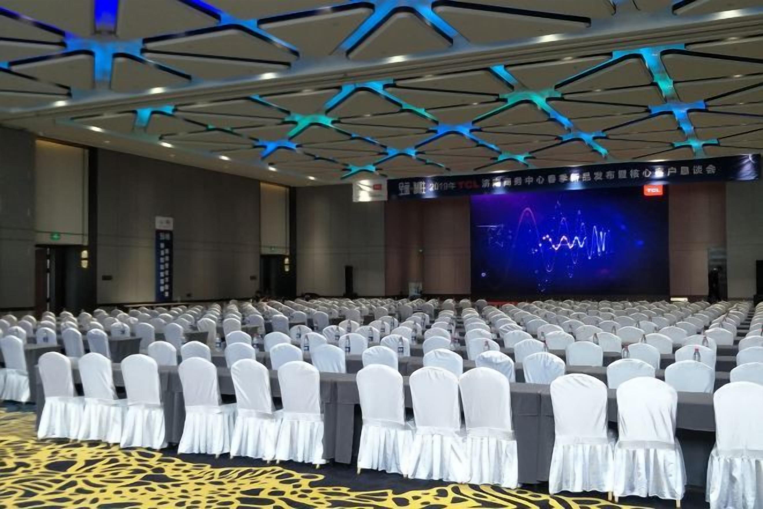 Une grande salle de conférence avec des rangées de chaises blanches, une scène dotée d’un écran et un design de plafond moderne.