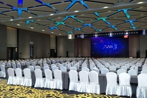 Ein großer Konferenzraum mit Reihen weißer Stühle, einer Bühne mit Bildschirm und einem modernen Deckenstil.