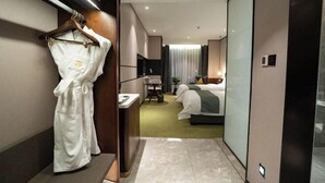 Habitación de hotel con bata colgada en un perchero, cama con manta verde, escritorio con silla y una pequeña mesa con una planta.