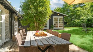 A wooden picnic table with wicker chairs, a small tree, and a house with a dark exterior and white windows.