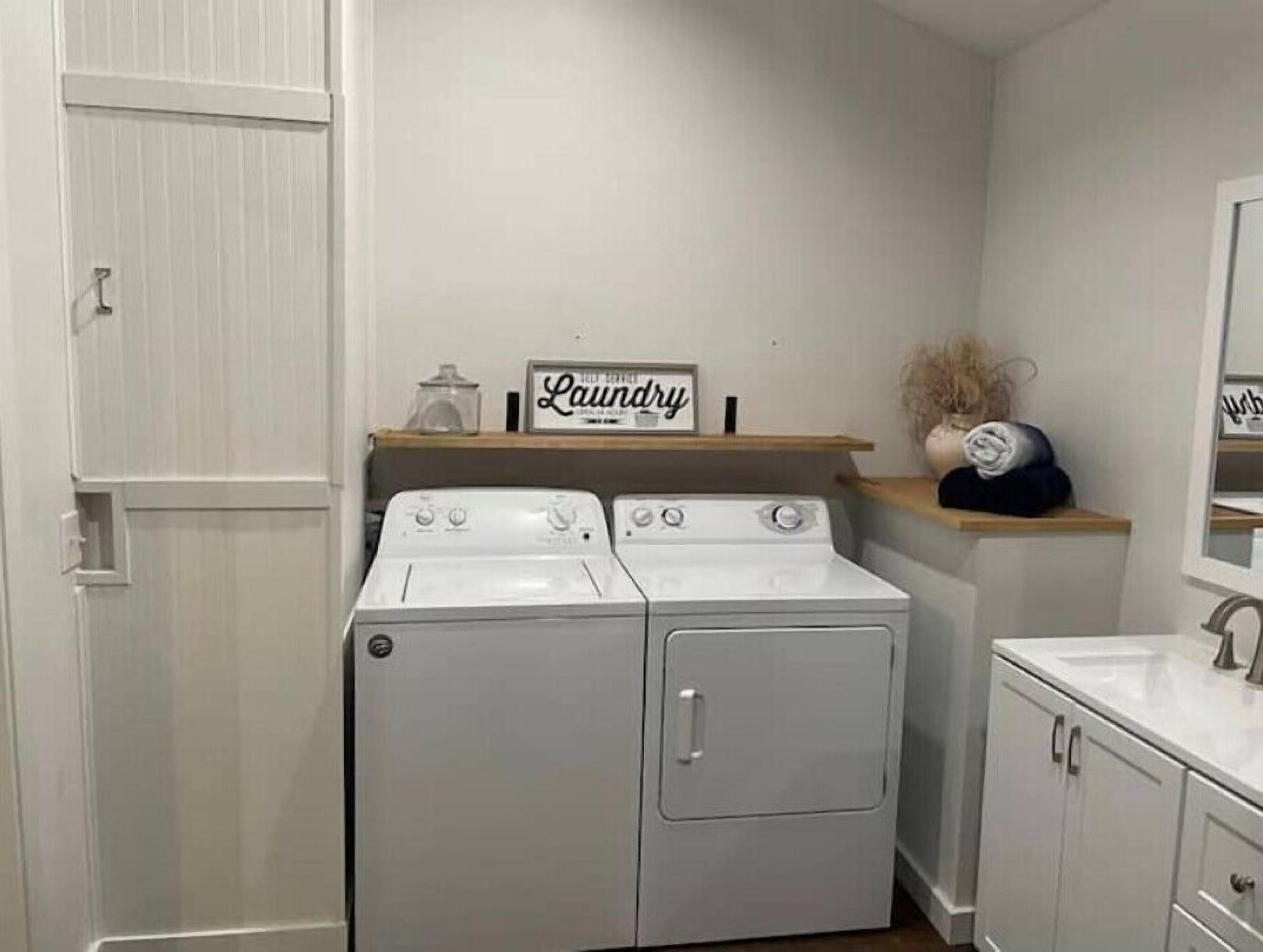 Un espace buanderie avec deux sèche-linge blancs, une étagère portant l’inscription « Buanderie », et un évier avec des placards.
