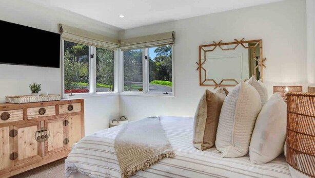 Une chambre à coucher avec un lit, une commode en bois, un grand miroir et une fenêtre donnant sur un paysage verdoyant.