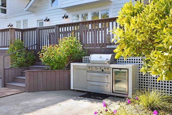 Un área de cocina al aire libre con parrilla, horno integrado y un banco para sentarse, rodeada por una terraza de madera y follaje.
