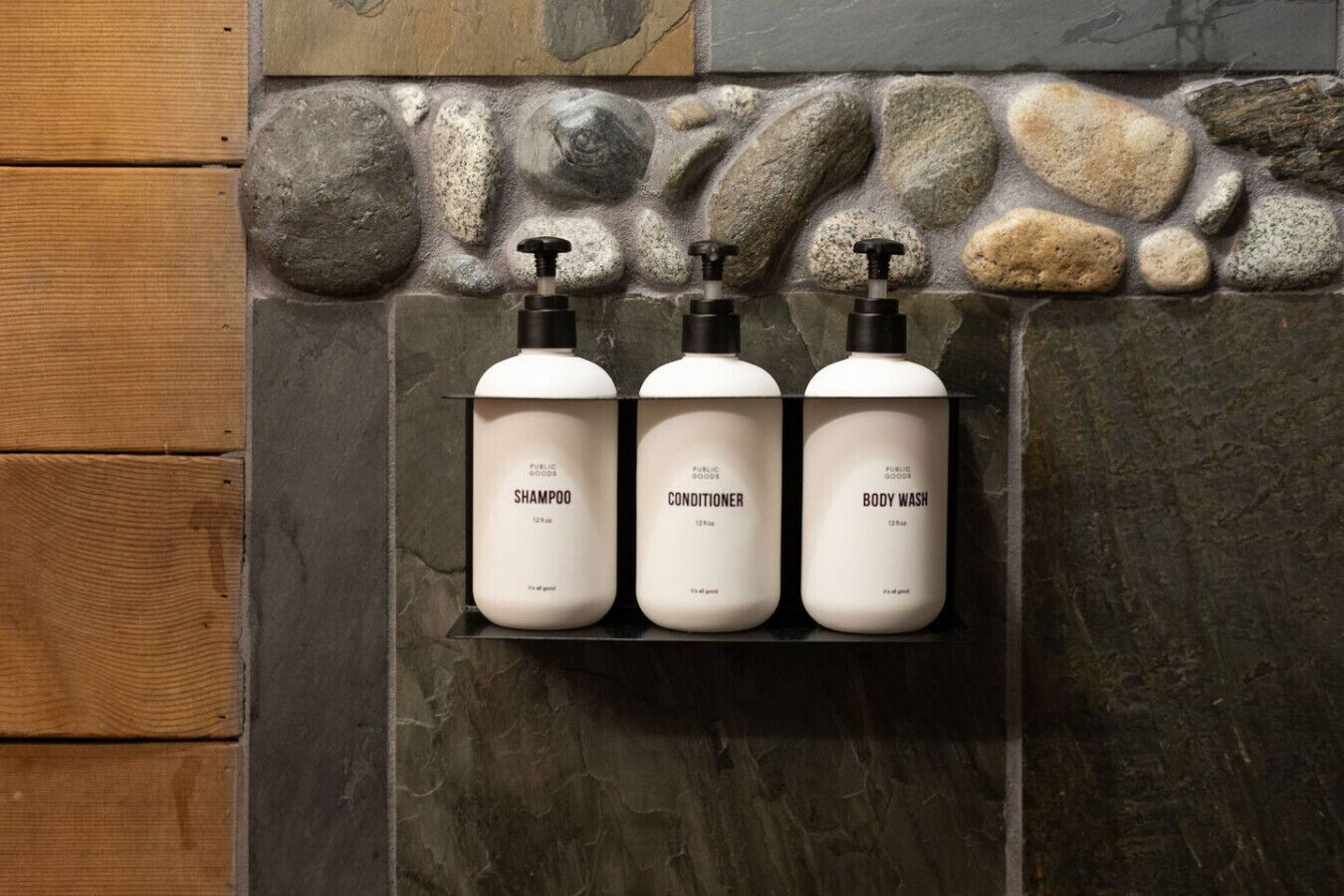 Drei Flaschen Shampoo, Conditioner und Duschgel auf einem Regal vor einer Steinwand.