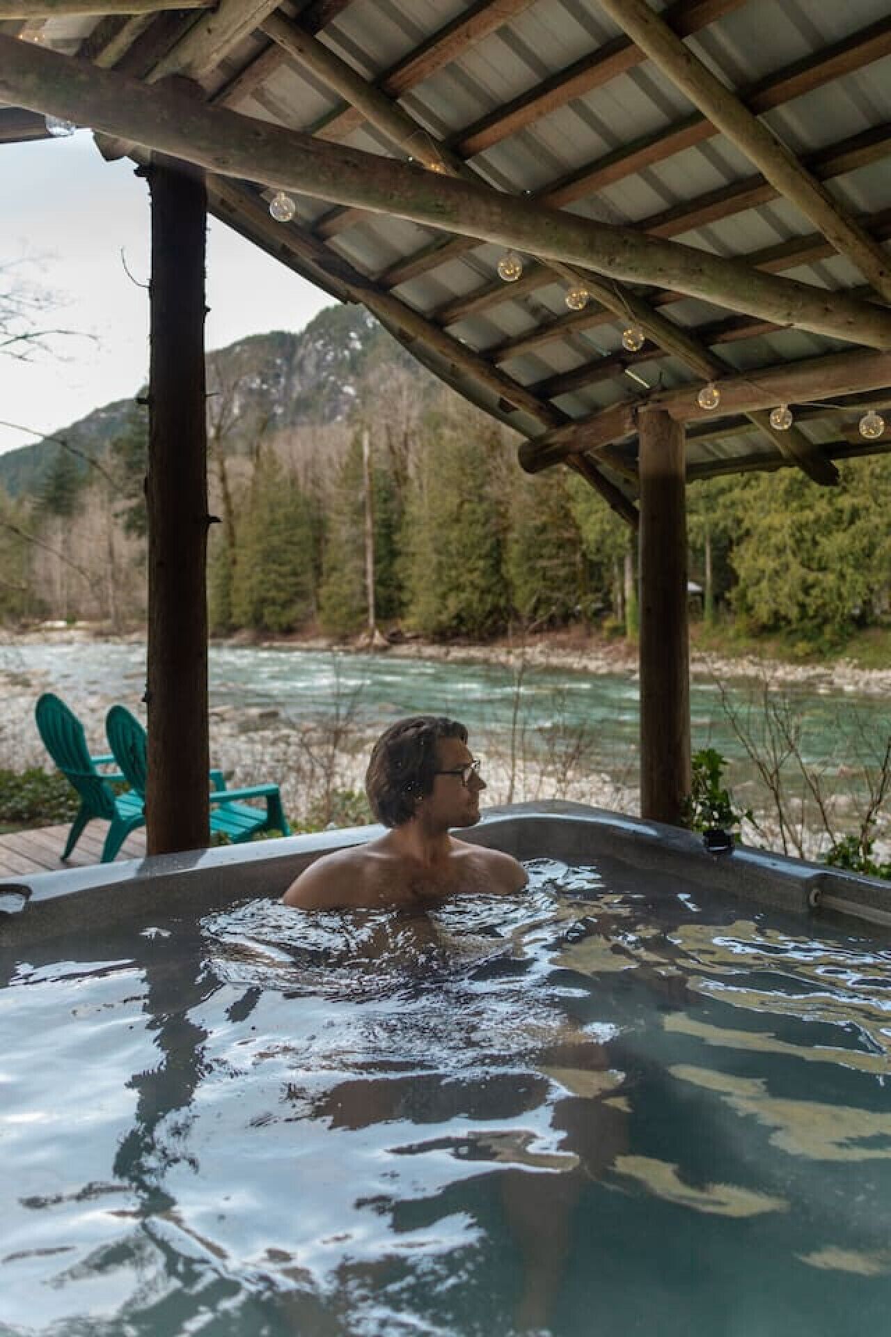 Eine Person in einer Whirlwanne mit Blick auf einen Fluss, Holzbalken und einem grünen Stuhl.