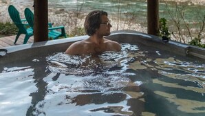 Eine Person in einer Whirlwanne mit Blick auf einen Fluss, Holzbalken und einem grünen Stuhl.