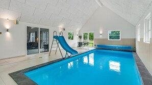 Une piscine intérieure avec un toboggan bleu, un espace détente et une porte vitrée donnant sur un espace extérieur.