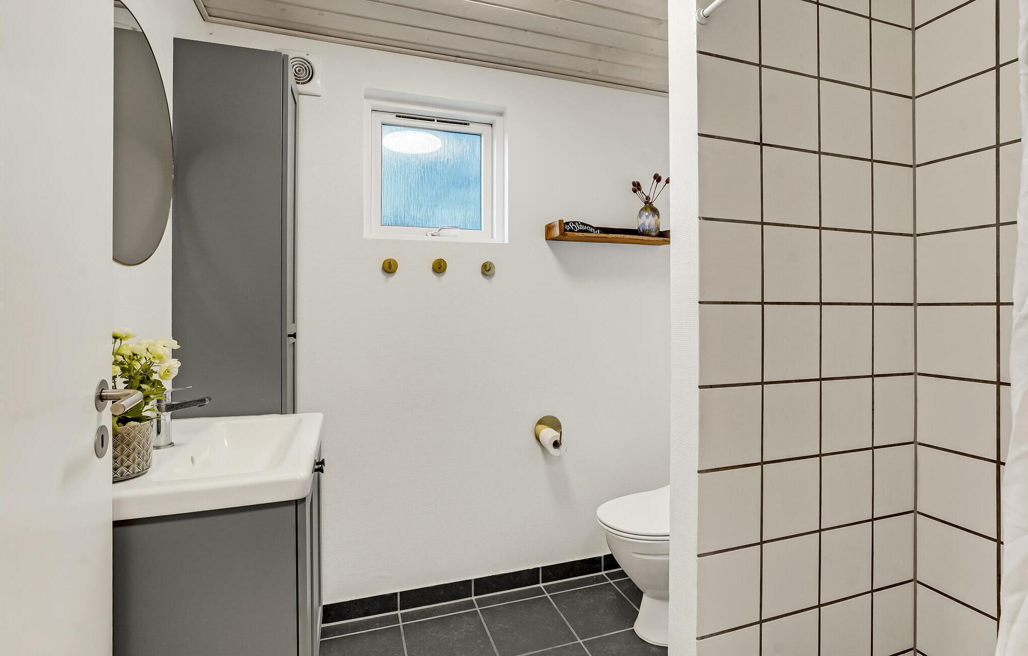 A bathroom with a white sink, a mirror, a window, a toilet, and a shower area with tiled walls.