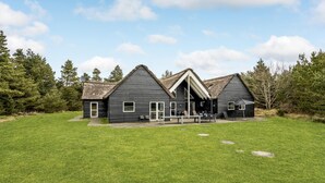 Une maison à deux étages avec un toit de chaume, une grande terrasse et une cour couverte d’herbe.