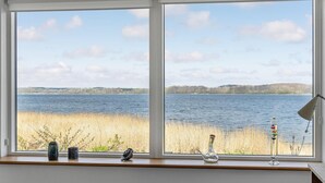 A room with a large window overlooking a lake, a wooden shelf with decorative items, and a lamp on the right. - 4 bedroom amazing home in Mariager (Mariager)