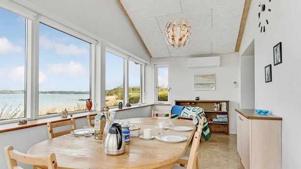 A dining area with a wooden table set for four, large windows offering a view of the sea, and a kitchen area with a wooden counter and a blue glass bowl. - 4 bedroom amazing home in Mariager (Mariager)