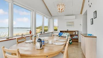 Spiseområde med et træbord dækket til fire personer, store vinduer med havudsigt, og et køkkenområde med en træbordplade samt en blå glasskål.
