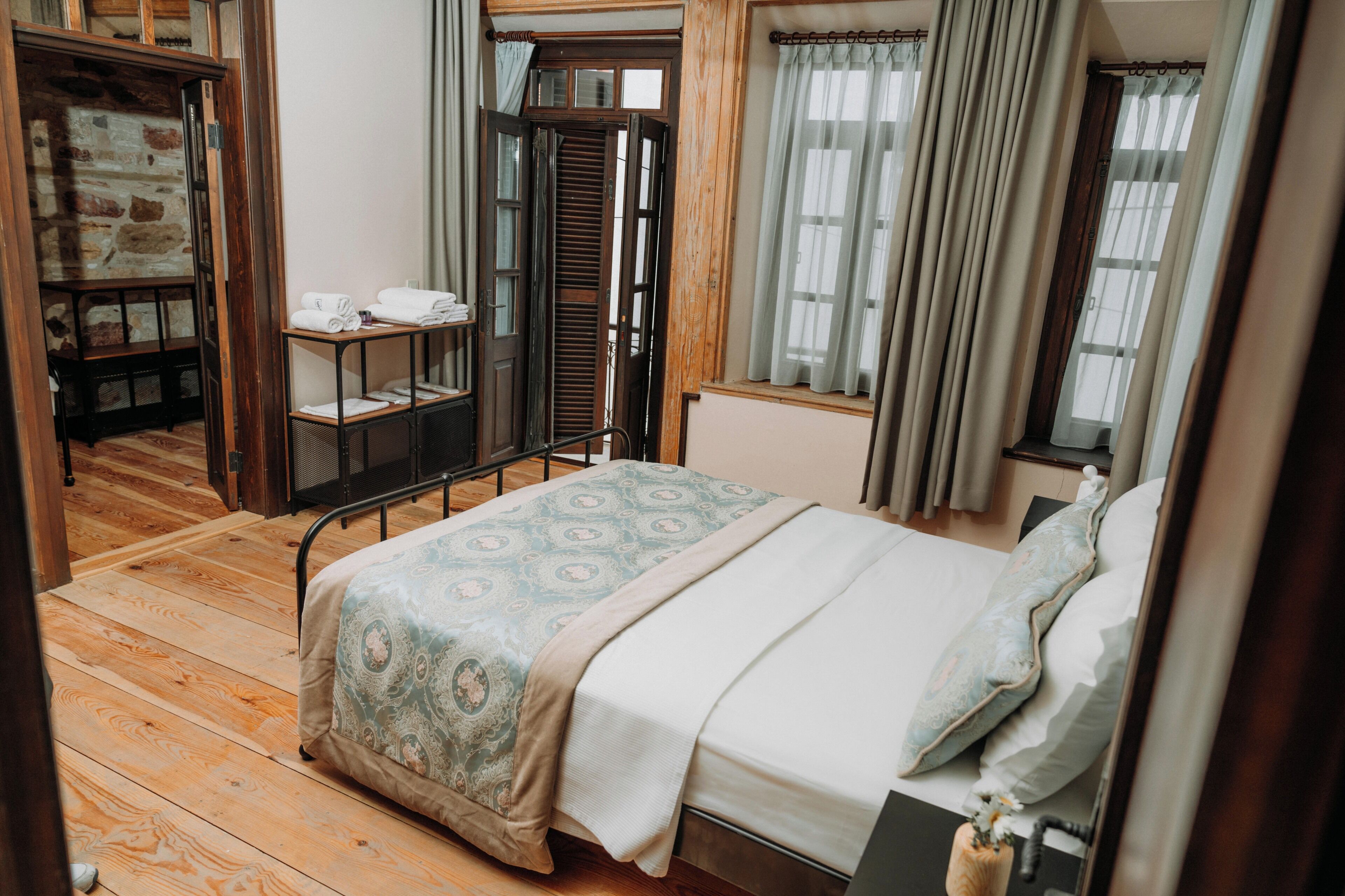 Un dormitorio con suelo de madera, una cama con ropa de cama estampada, una mesita de noche, una ventana con cortinas y una puerta que da a otra habitación.