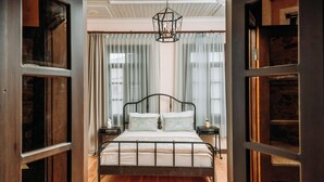 A bedroom with a wooden floor, a metal bed frame, a hanging light fixture, and curtains on the windows. - Nill's Stone House Otel (Cesme)