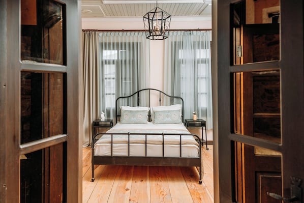 A bedroom with a wooden floor, a metal bed frame, a hanging light fixture, and curtains on the windows. - Nill's Stone House Otel (Cesme)