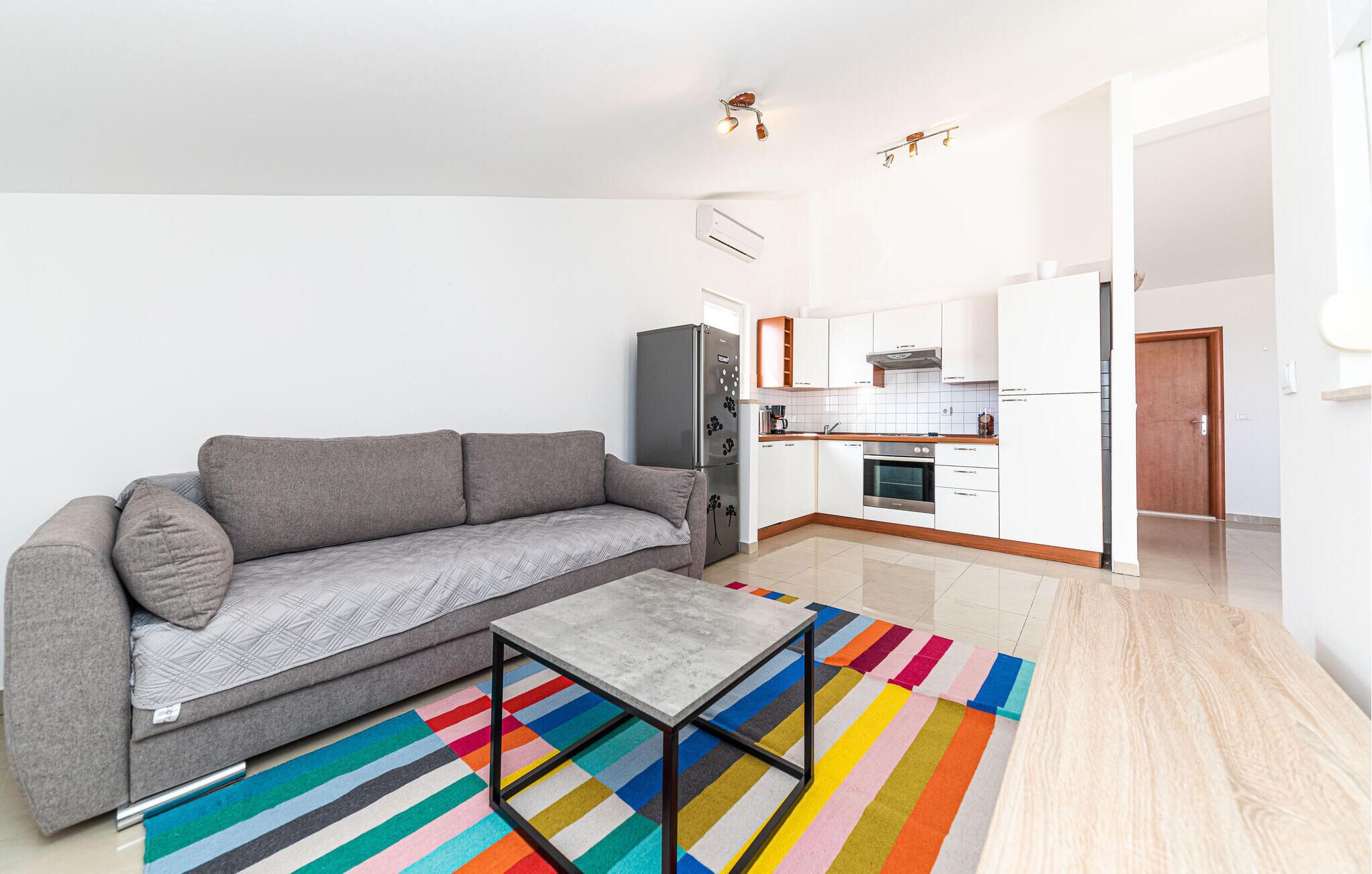 A compact living space with a grey sofa, a small table, a colorful striped rug, a kitchen with white cabinets and a wooden countertop, and a door leading to another room.