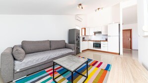 Ein kompakter Wohnraum mit grauem Sofa, kleinem Tisch, farbenfroher Streifenoptik-Teppich, einer Küche mit weißen Schränken und einer Holzarbeitsplatte sowie einer Tür zu einem weiteren Zimmer.