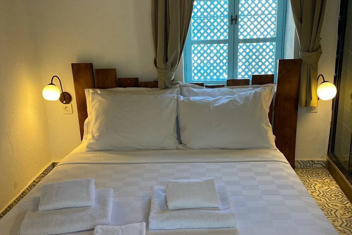 A neatly made bed with pillows and folded towels, a window with curtains, and wall-mounted lights.