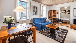 A modern living room with a blue sofa, wooden coffee table, and a dining area with a wooden table and chairs. There is a bookshelf, a radiator, and a window with curtains.