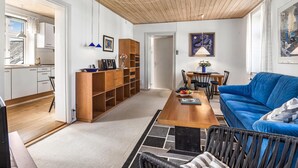 A modern living room with a blue sofa, a wooden coffee table, and a dining area with a table and chairs. There is a kitchen visible in the background with white cabinets and a wooden ceiling.