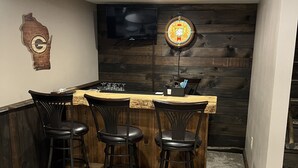 A bar area with a wooden counter, four black bar stools, a television mounted on the wall, and a decorative wall piece. - Up North, Outdoor Enthusiast’s Paradise! (Birchwood)